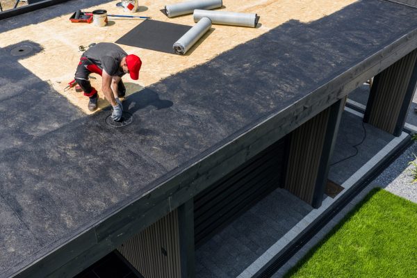 A construction worker is installing EPDM roofing material on a contemporary home under clear skies, surrounded by tools and prepared materials for the roofing project.